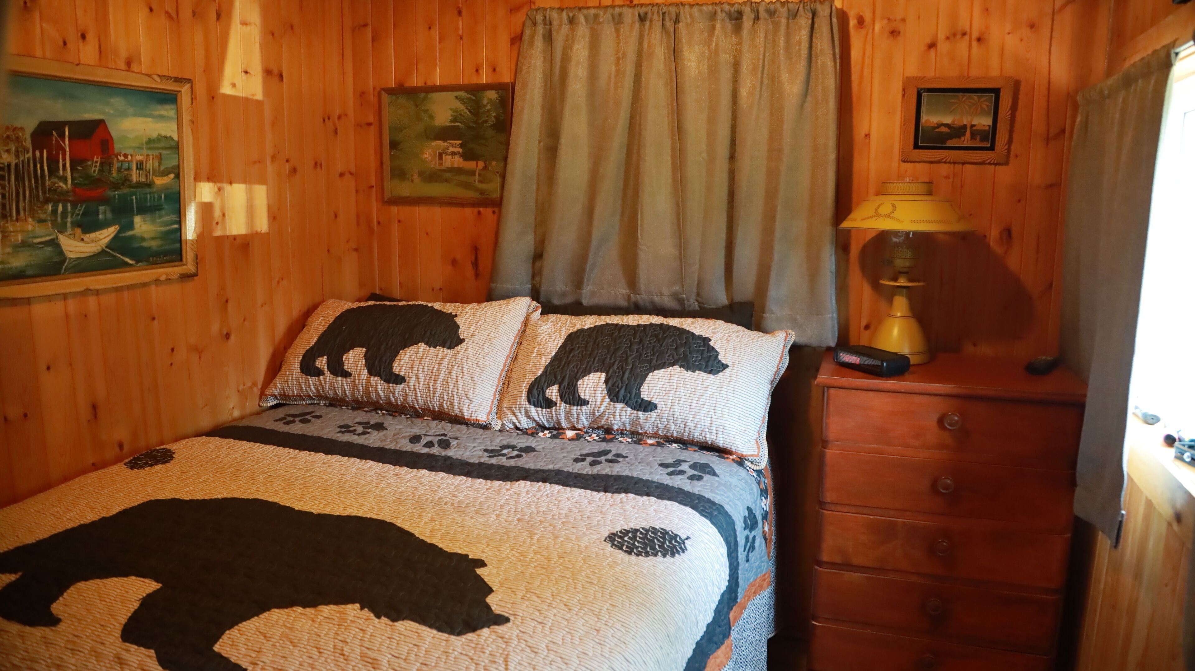 Une pièce aux murs lambrissés, avec un lit recouvert d’une courtepointe à motifs d’ours, une table de chevet avec une lampe et deux tableaux encadrés accrochés au mur.