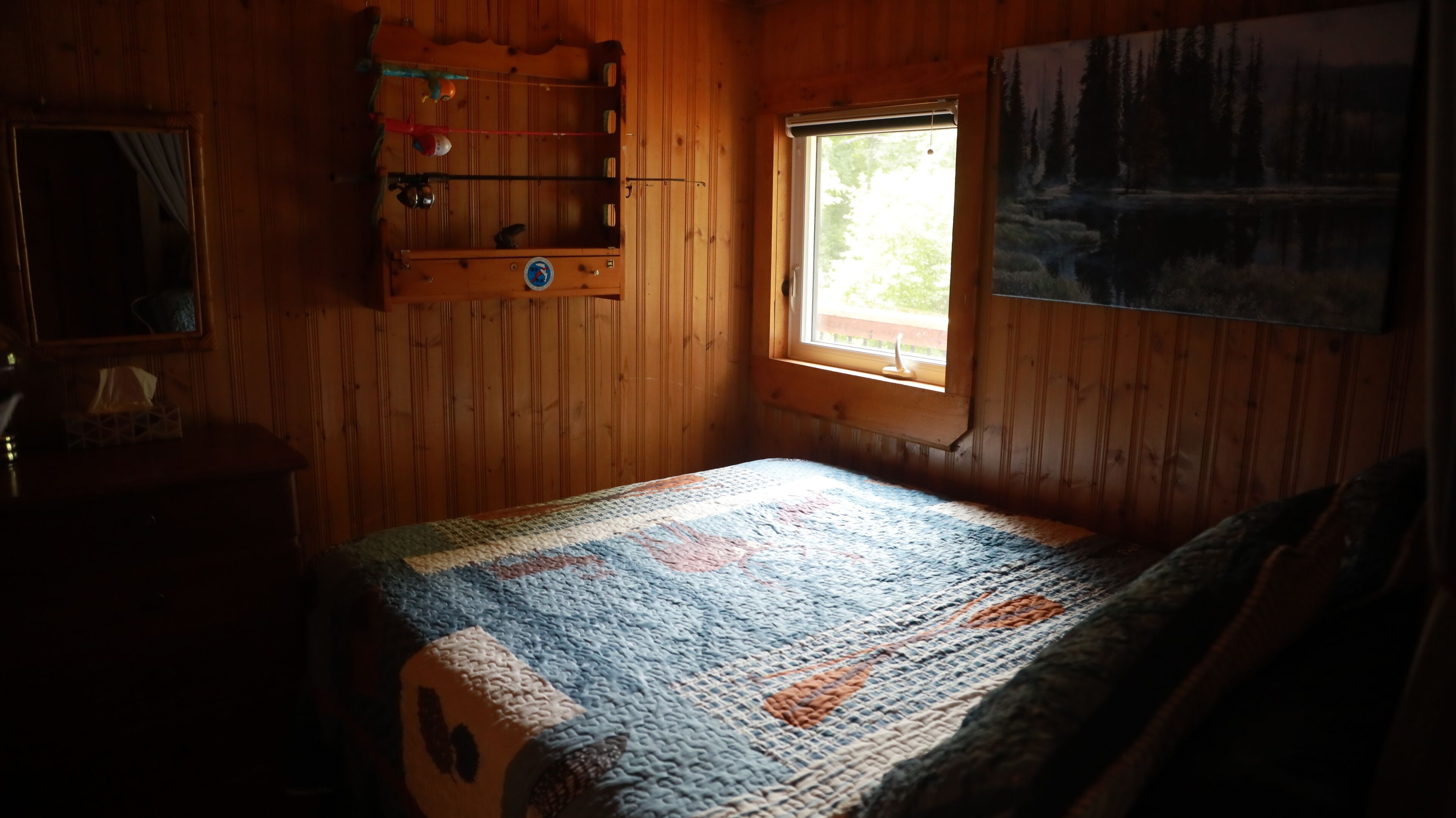 A wooden room with a bed, a window, a shelf with books, and a painting.