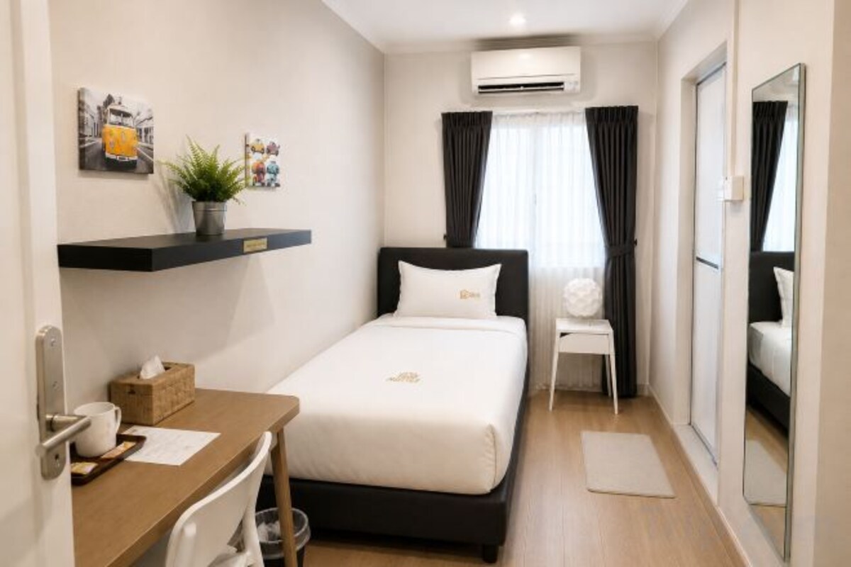 A hotel room with a bed, a desk, a chair, a small table, a potted plant, a mirror, and a window with curtains.