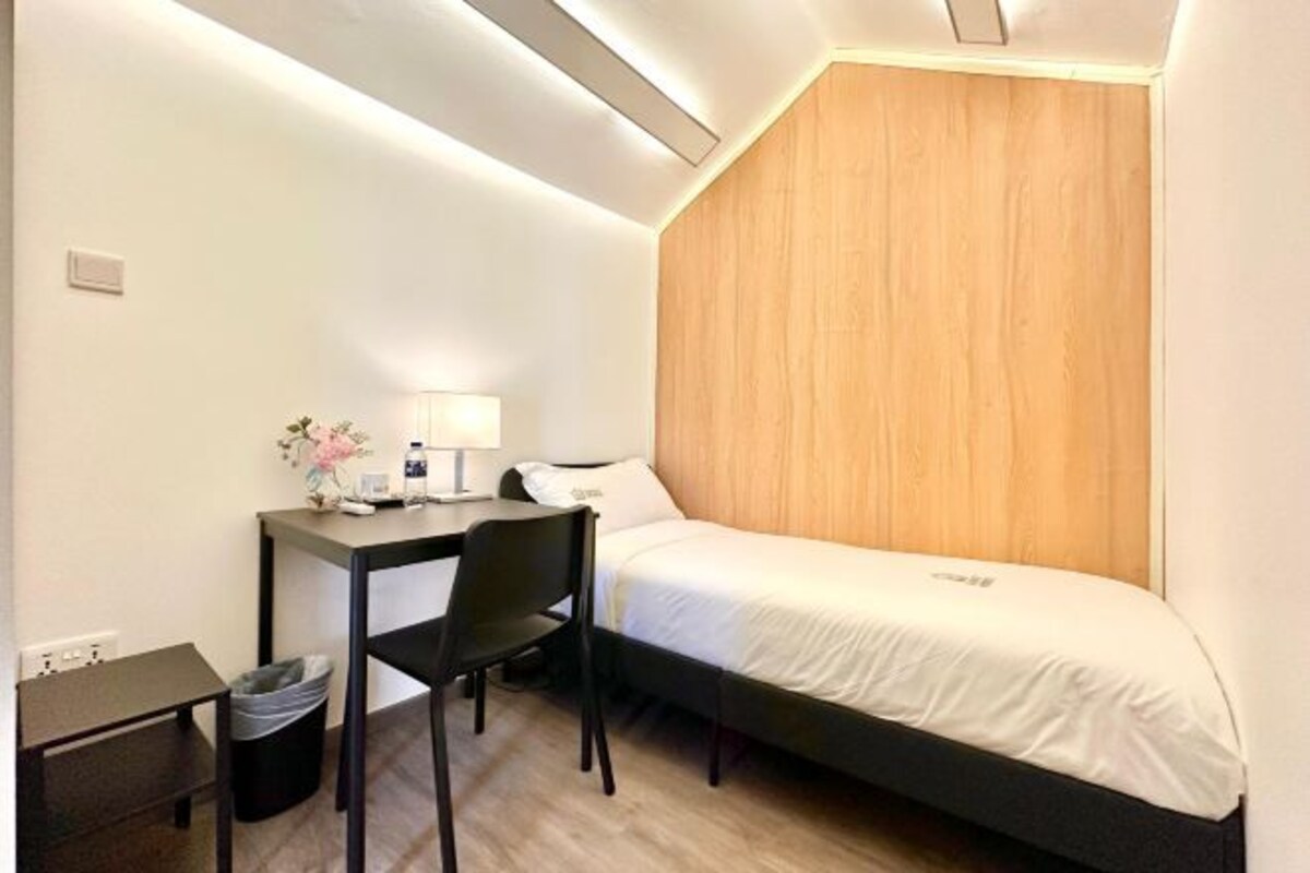 Habitació d'hotel petita amb un llit individual, una escriptori amb cadira, una taula petita i un gresol.