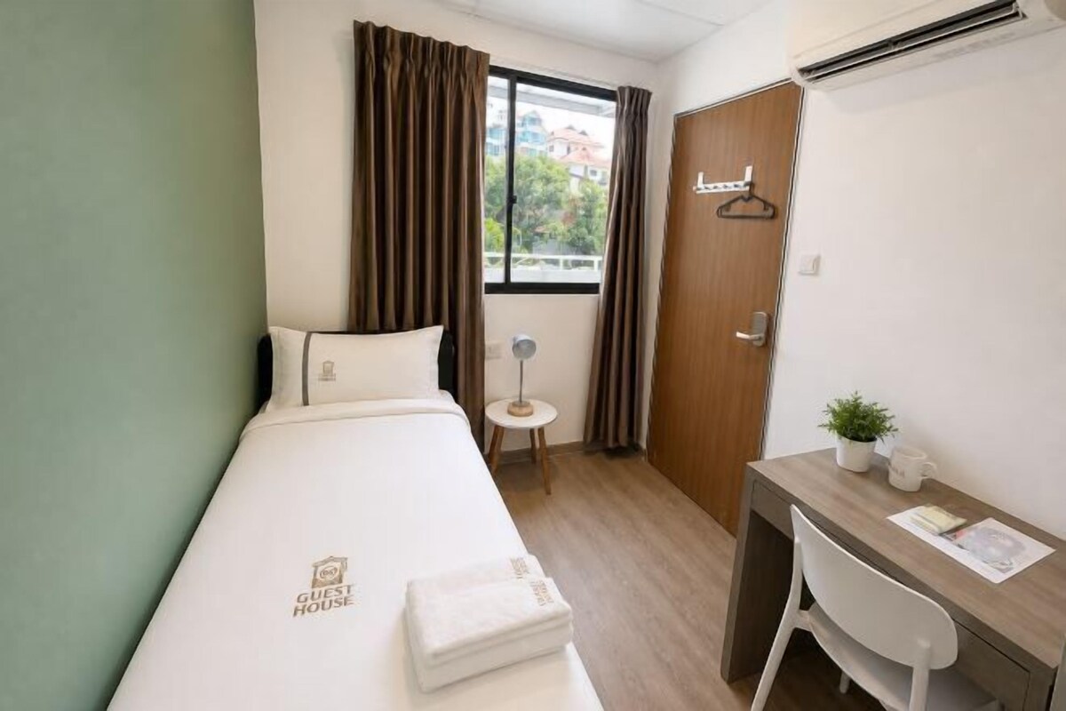 A single bed with a 'Guest House' logo, a desk with a chair, a small table with a lamp, and a window with curtains.