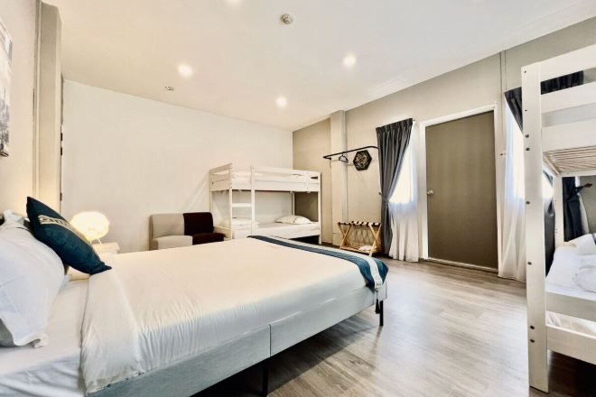 Habitació d'hotel moderna amb un llit gran, un altre llit més petit, una taula de treball amb una làmpada, un armari i una llitera.
