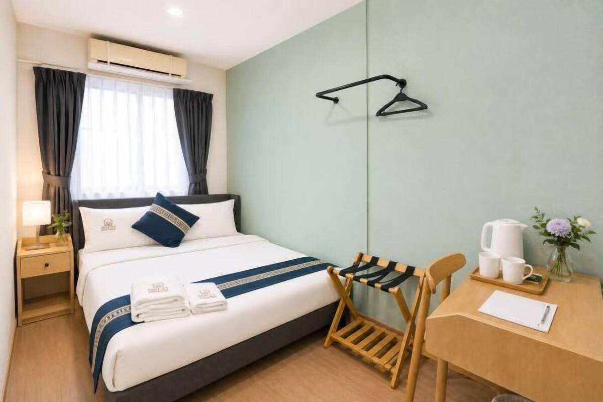 A hotel room with a bed, a desk with a chair, a hanging coat rack, and a small table with a kettle and cups.