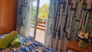 A room with a bed, a wooden nightstand, a window with patterned curtains, and a view of a balcony with a railing.