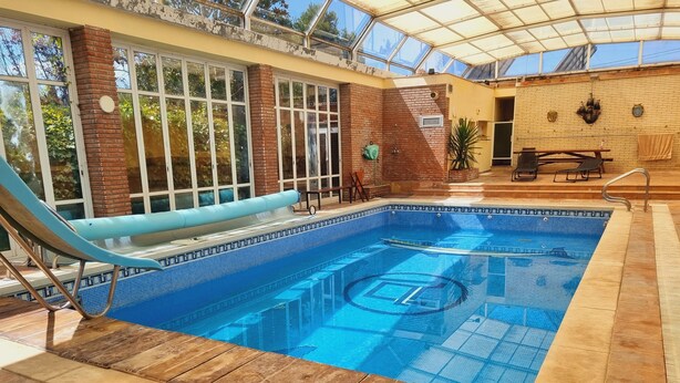 An indoor swimming pool with a blue tiled floor, a lounge chair, and a glass-enclosed roof.