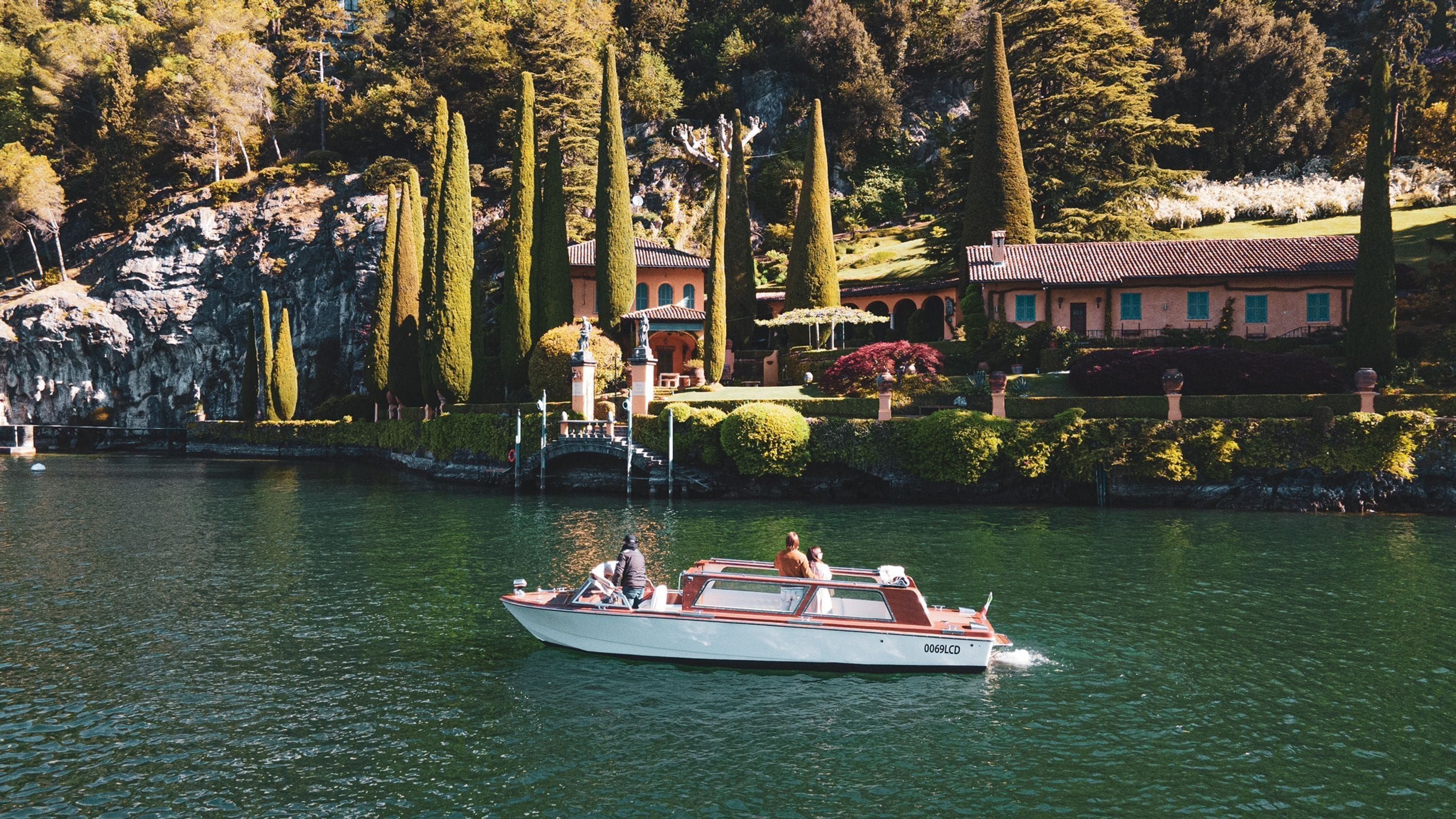A boat on a calm lake with a rocky shoreline and a building with a red roof in the background.