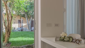 Ein Zimmer mit weißem Schreibtisch, einem kleinen Tisch mit Blumen und Blick auf eine grüne Fläche mit Bäumen und einem Gebäude im Hintergrund.