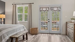 Ein Schlafzimmer mit einem Bett, einer Bank, einem Korbwäscher, einem Schminktisch und einer Tür zu einem Außenbereich mit Stuhl und Tisch.