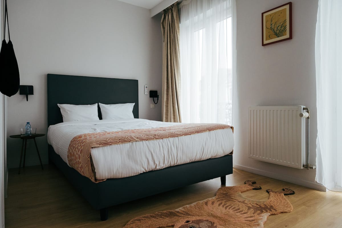 A neatly arranged bedroom with a bed, bedside tables, a radiator, and a window with curtains.