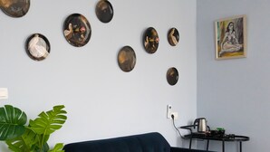 A living room with a dark blue sofa, a round coffee table, and a plant. There are wall-mounted decorative plates and a painting. - Hôtel La Pièce  (Brussels)