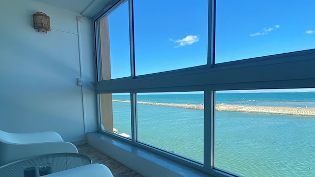 A room with a large window overlooking a beach and water. There are two white chairs and a small table with two cups.
