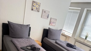 Camera da letto moderna con due letti singoli, un comodino in legno, una sedia grigia e una finestra con persiane.