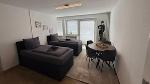 Un ambiente moderno e funzionale con un letto, un divano, un tavolo da pranzo con sedie e un'ampia finestra con tende.