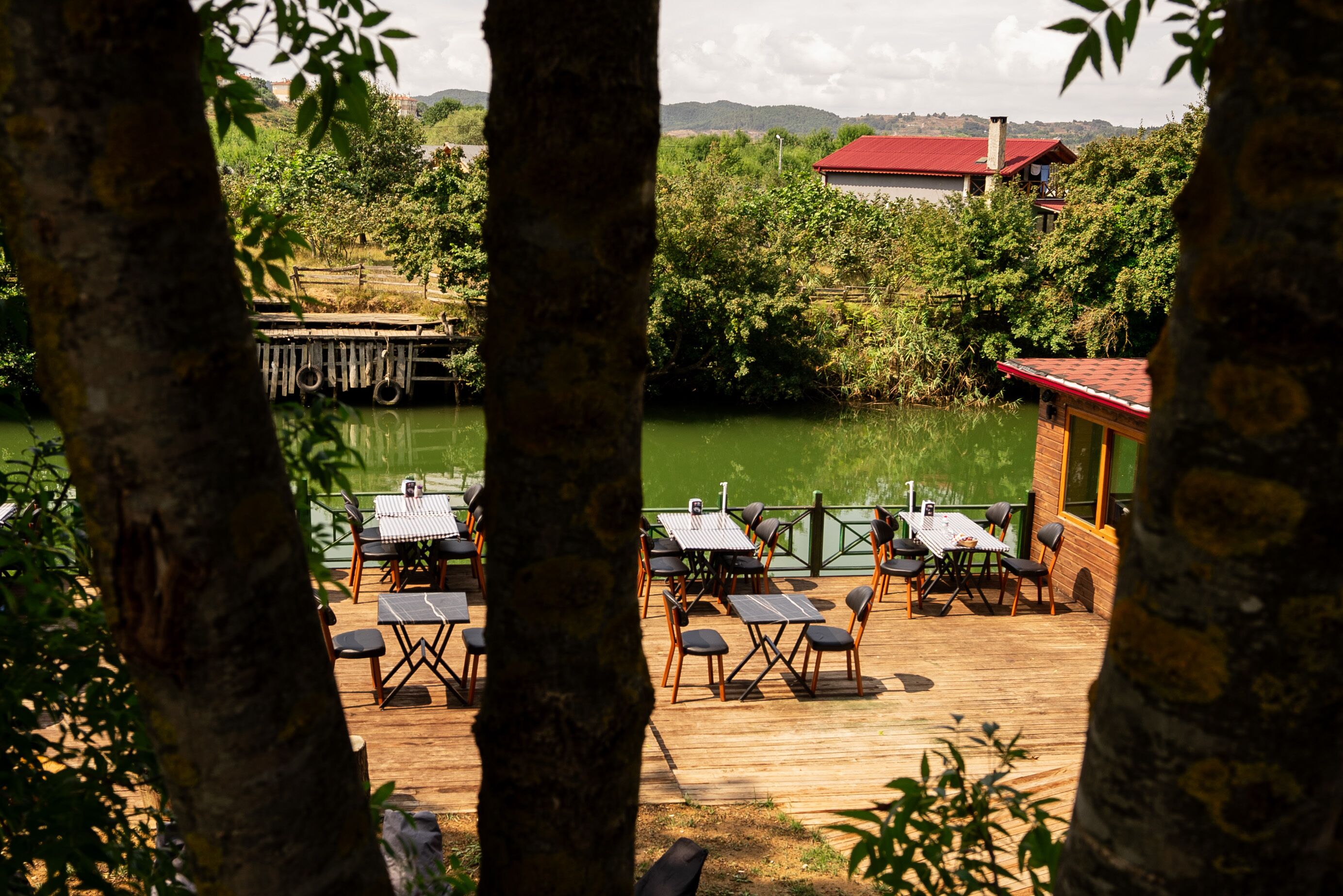 An outdoor dining area with tables and chairs, a wooden deck, and a view of a river and greenery.