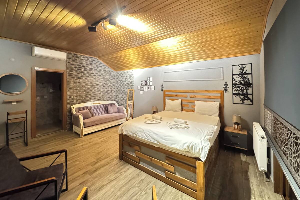 A bedroom with a wooden ceiling, a stone wall, a bed, a sofa, and a mirror.