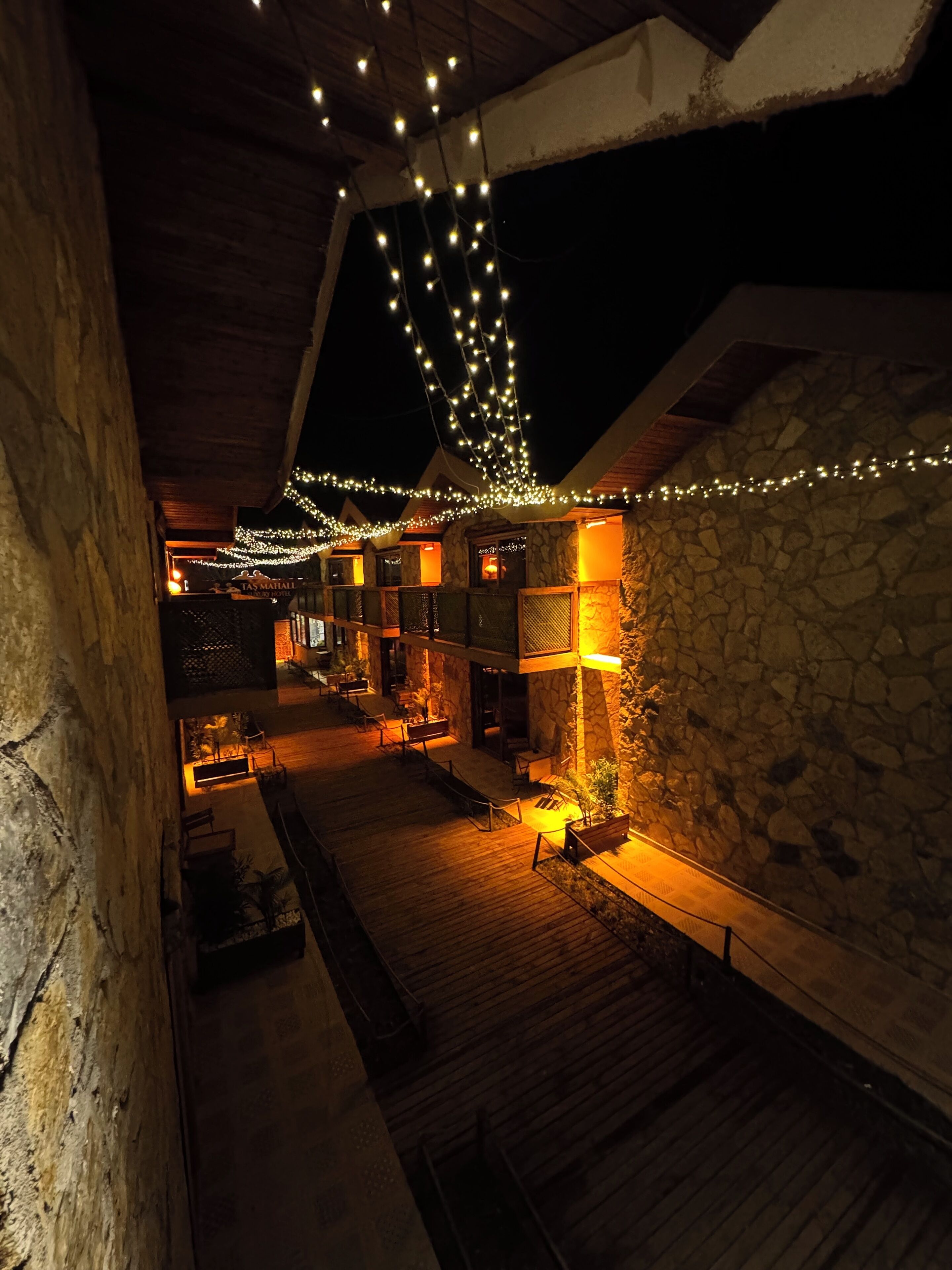 Un área de estar exterior acogedora y con poca luz, con paredes de piedra y suelo de madera, decorada con luces colgantes.