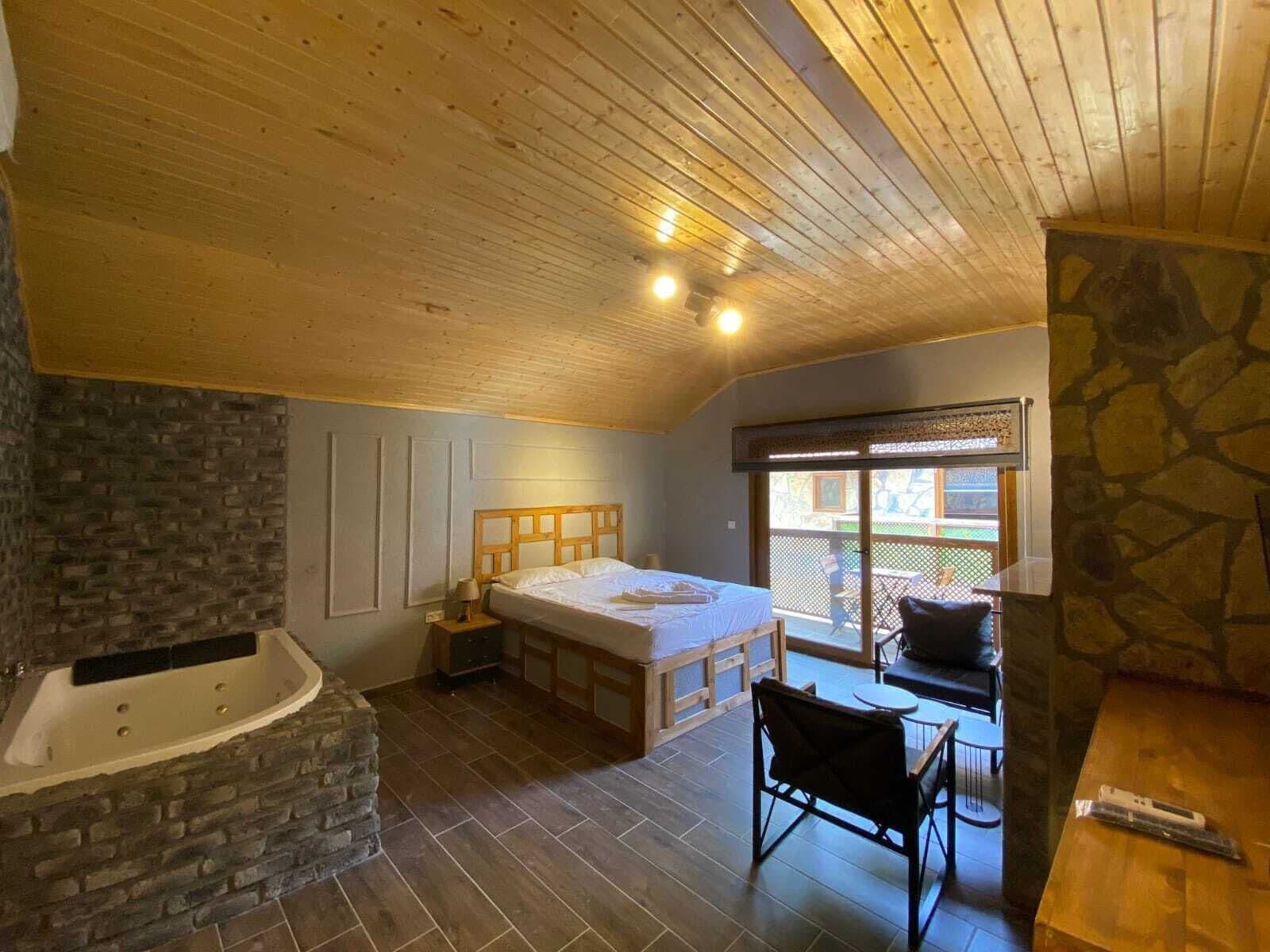 Un dormitorio con techo de madera, pared de piedra, una cama grande, una bañera y vistas al exterior.