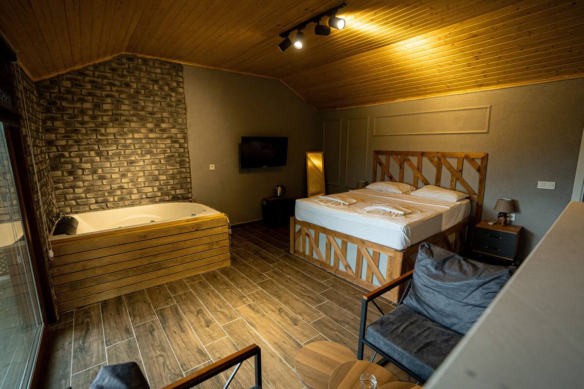 A room with a wooden ceiling, a brick wall, a large bed, a wooden bathtub, a television, and a small table with a lamp.