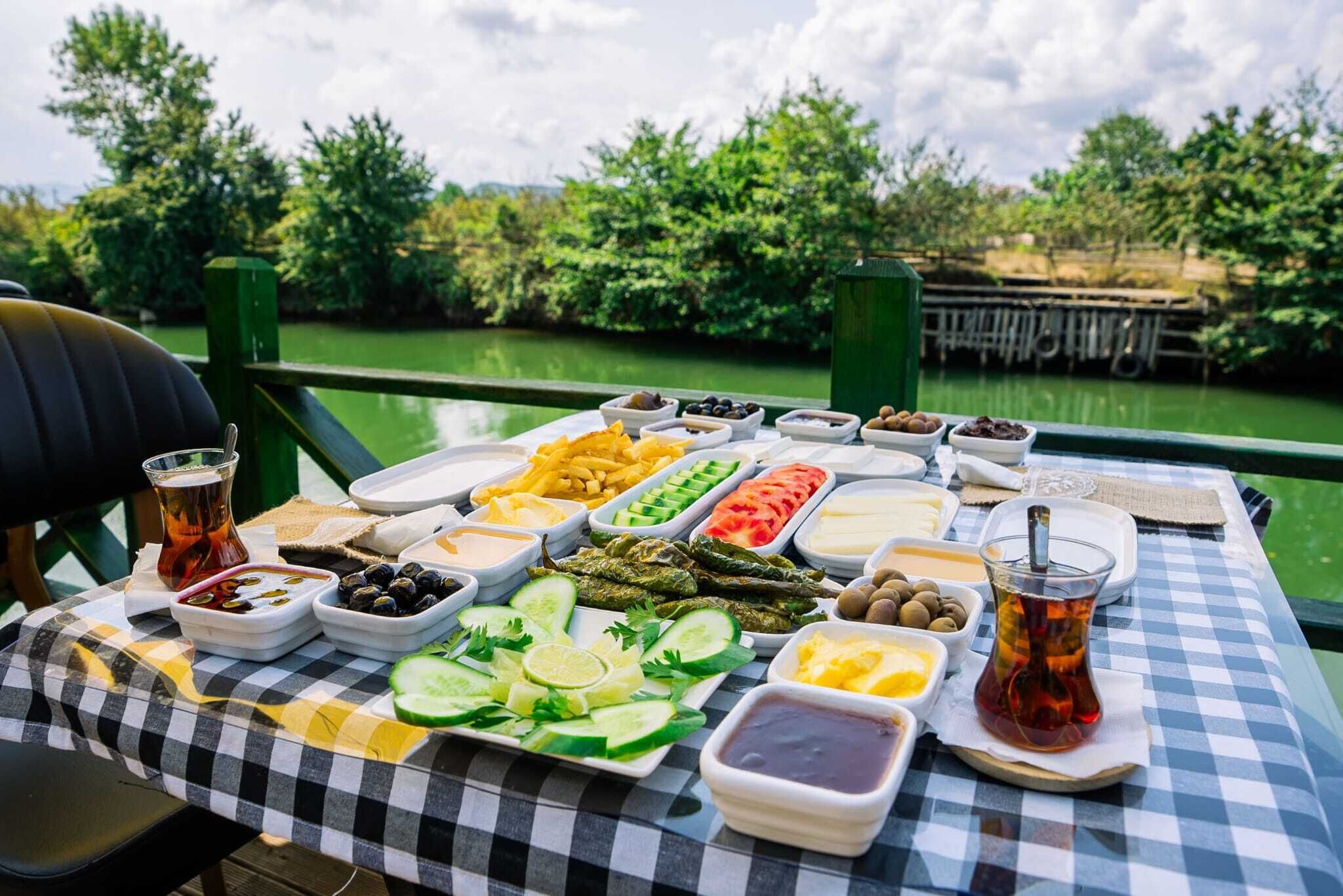 A table with various food items and drinks, set against a backdrop of greenery and a body of water.