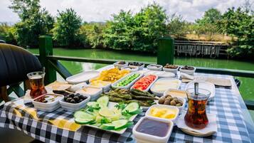 A table with various food items and drinks, set against a backdrop of greenery and a body of water.