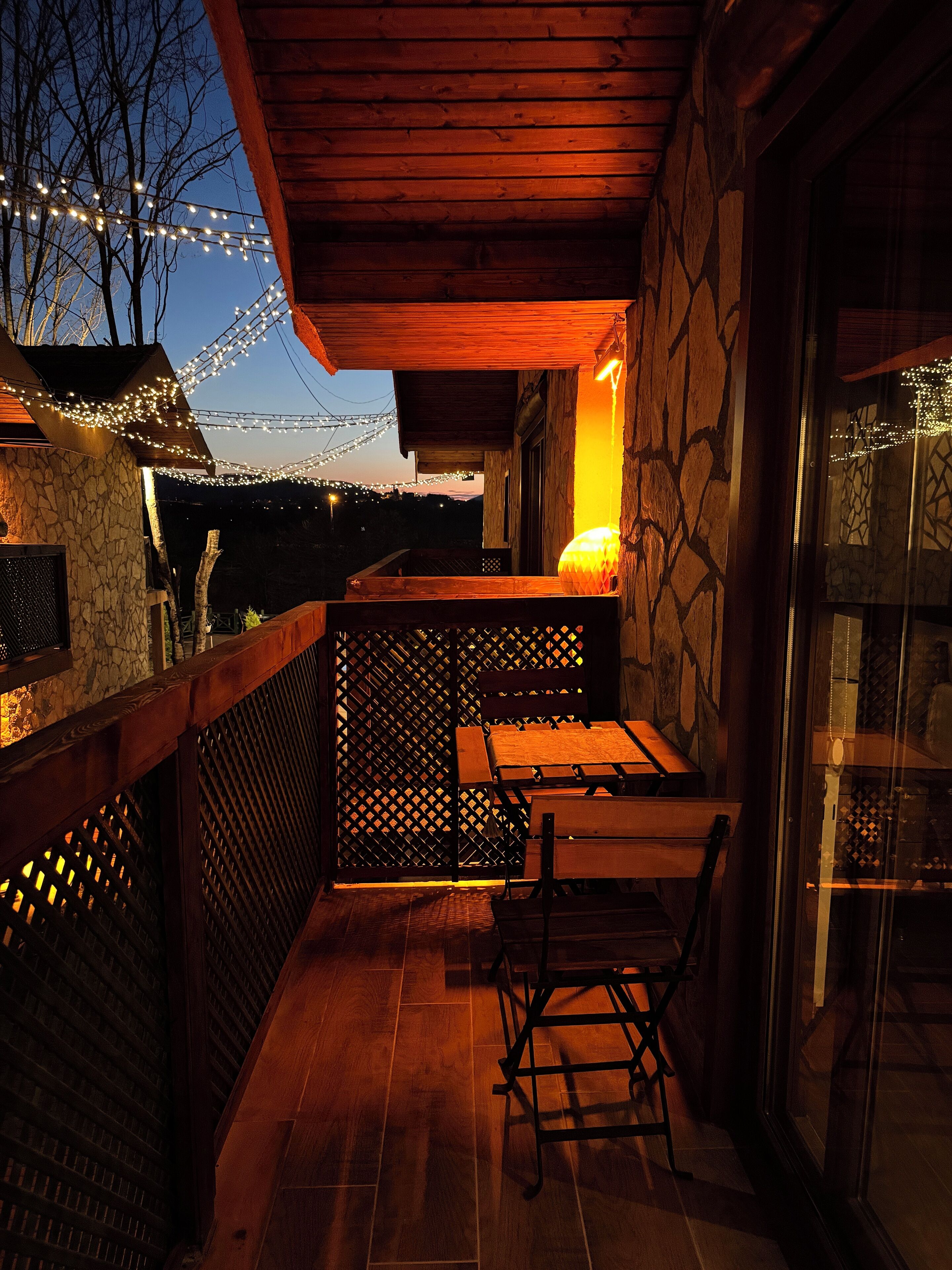 A wooden balcony with stone walls, string lights, and a table with chairs.