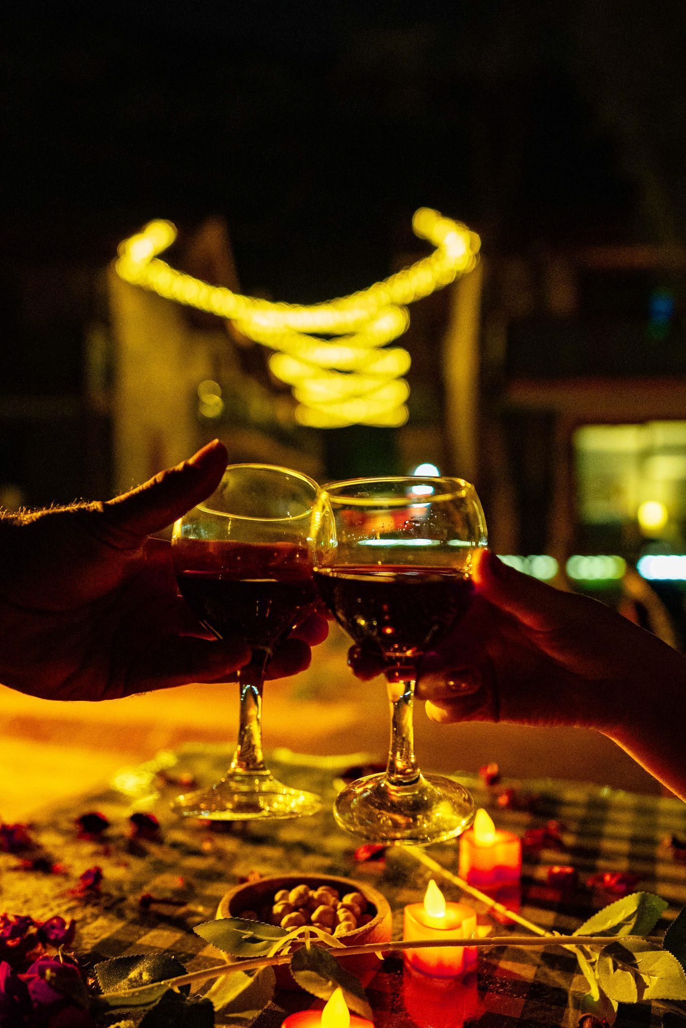 Deux mains tenant des verres à vin, une bougie et un bol de noix sur une table recouverte d’une nappe à carreaux.
