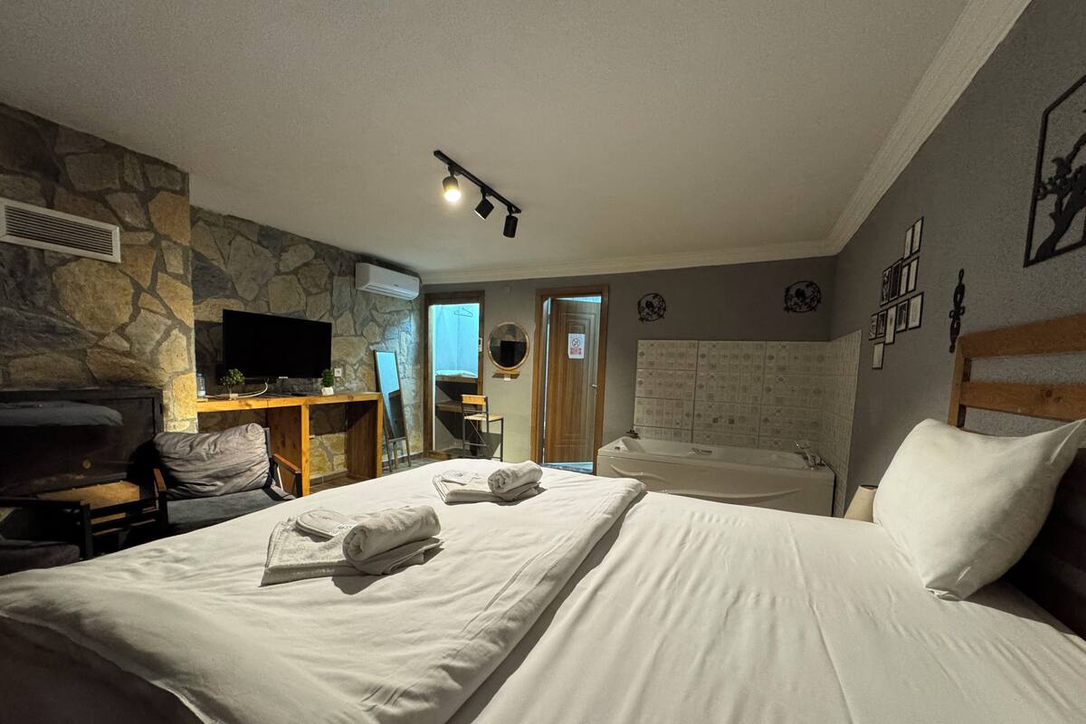 A hotel room with a stone wall, a flat-screen TV, a desk with a chair, a bed with white linens, and a wooden headboard.