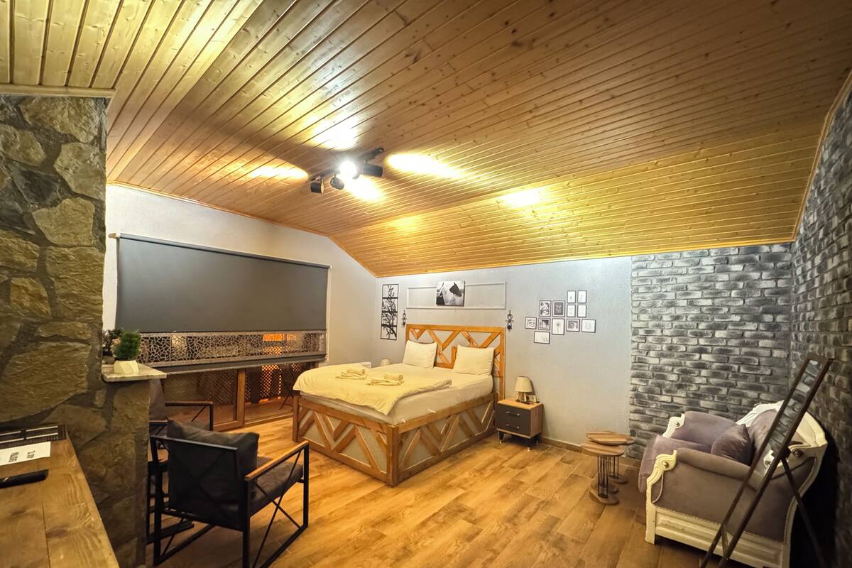 A cozy bedroom with a wooden ceiling, a large bed, a desk, and a stone wall.