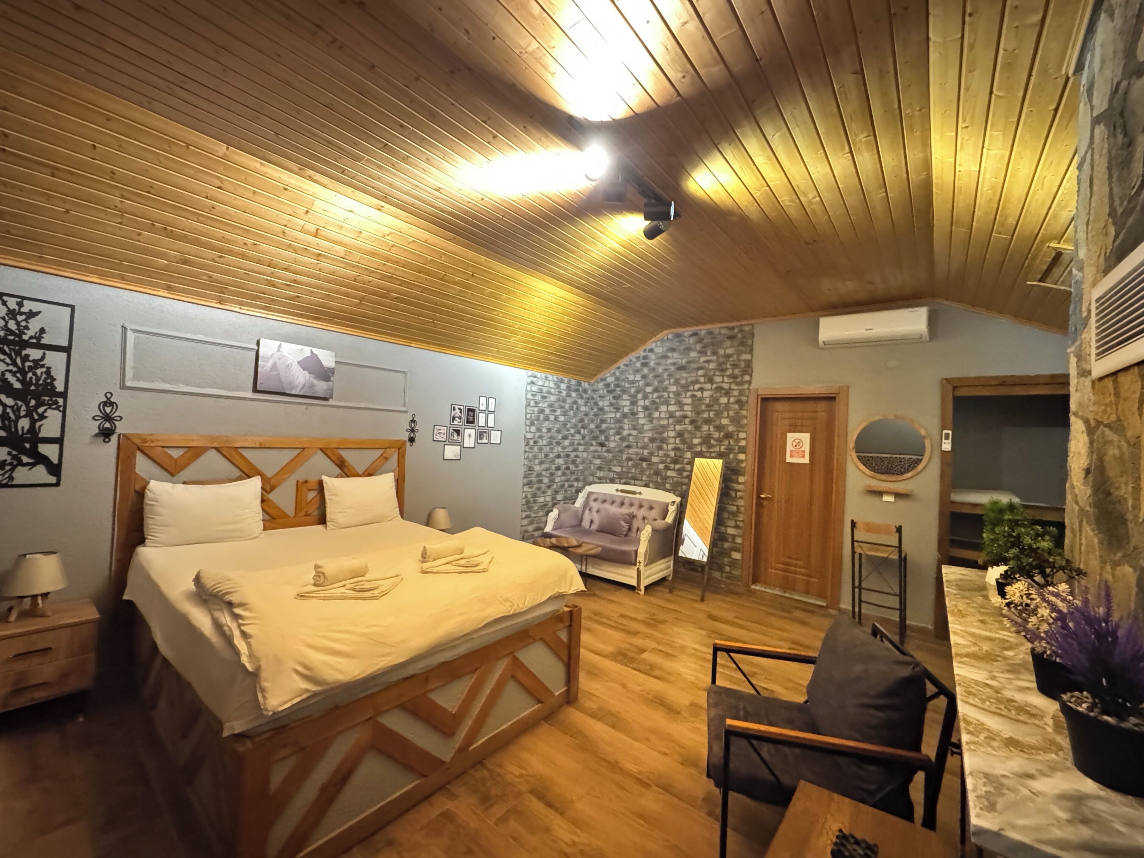 Ein gemütliches Schlafzimmer mit Holzboden, einem Bett mit beigefarbener Bettwäsche, einem Nachttisch, einer Kommode, einer Wand mit Steinoptik, einem Spiegel und einem Blumentopf.
