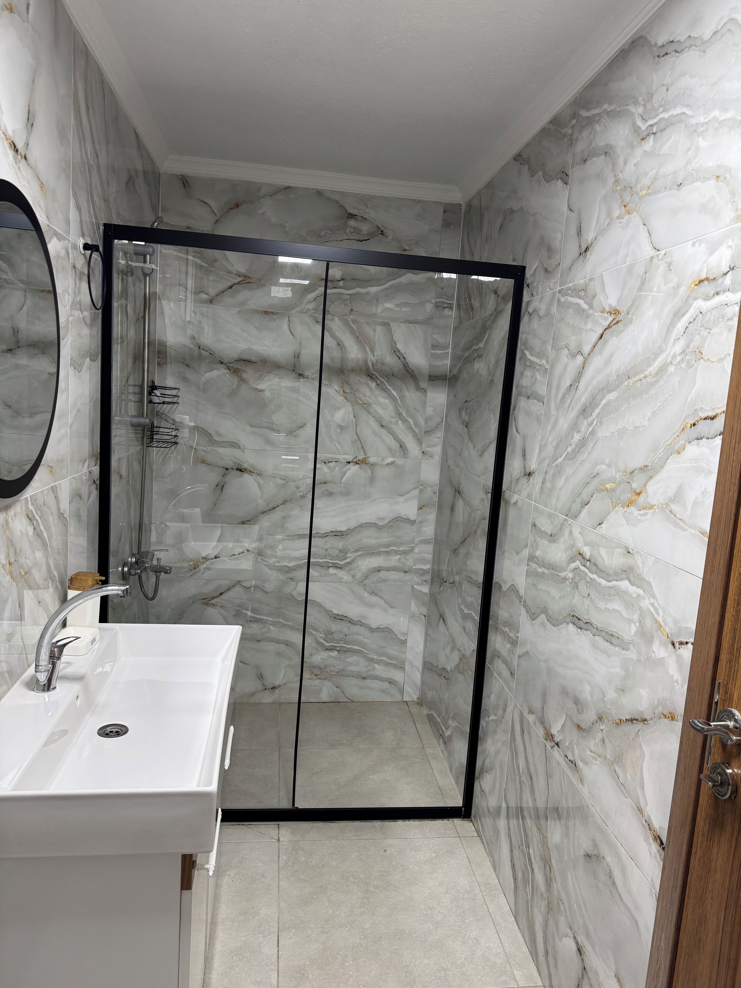 Un baño con ducha envuelta en vidrio, un lavabo blanco y paredes con diseño de mármol.