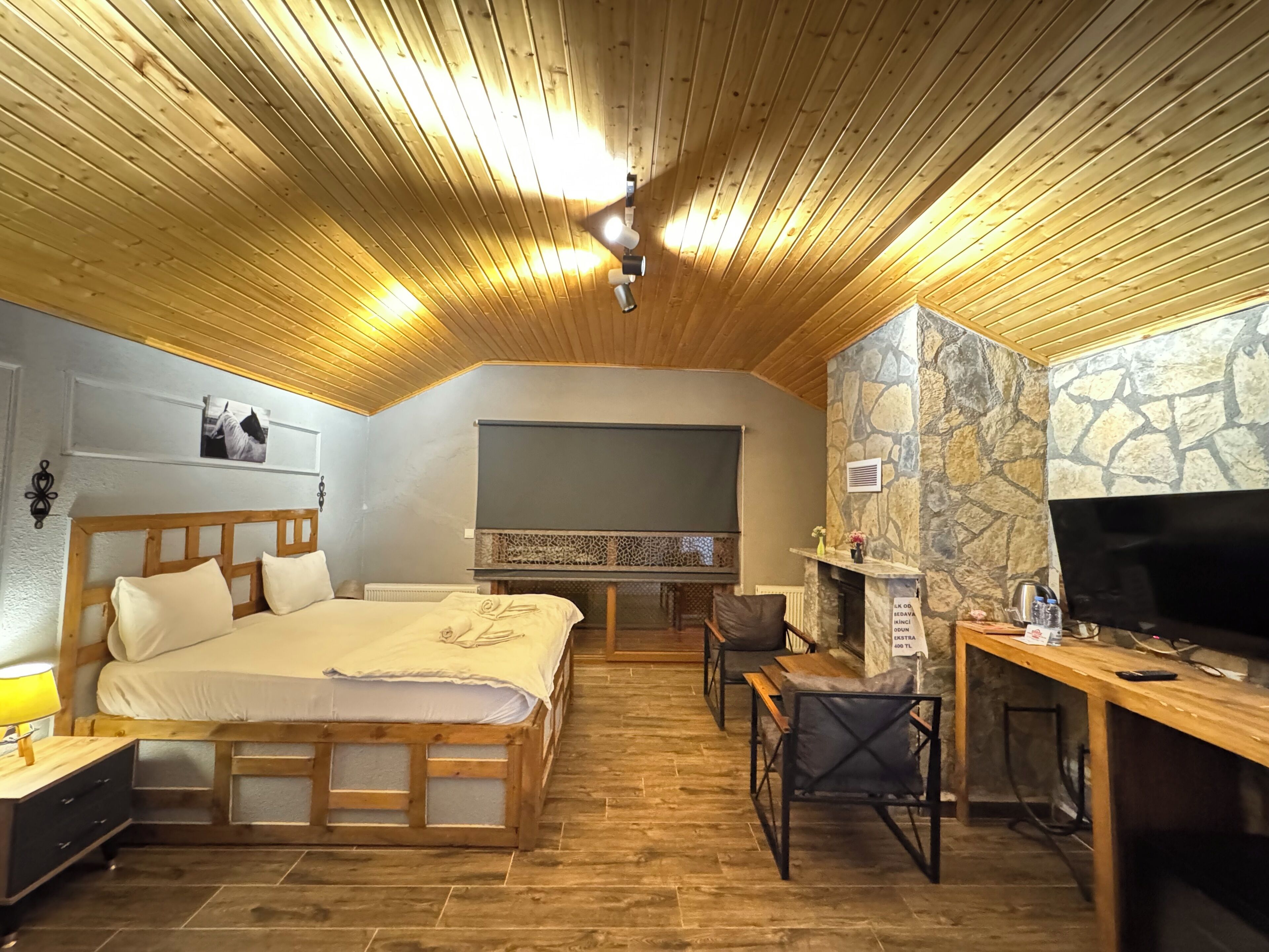 A cozy room with a wooden ceiling, a bed with a headboard, a desk with a chair, and a stone wall.