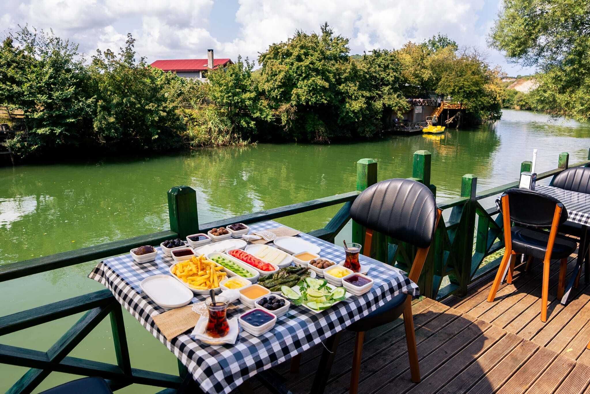 A dining table with a variety of food items and drinks on a checkered tablecloth, overlooking a river with greenery and a building in the background.