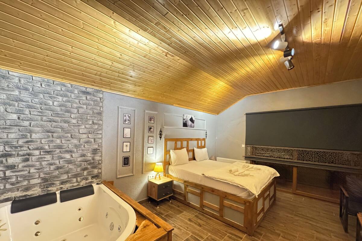 A bedroom with a wooden ceiling, a stone wall, a large bed, a wooden cabinet, a bathtub, and a TV.