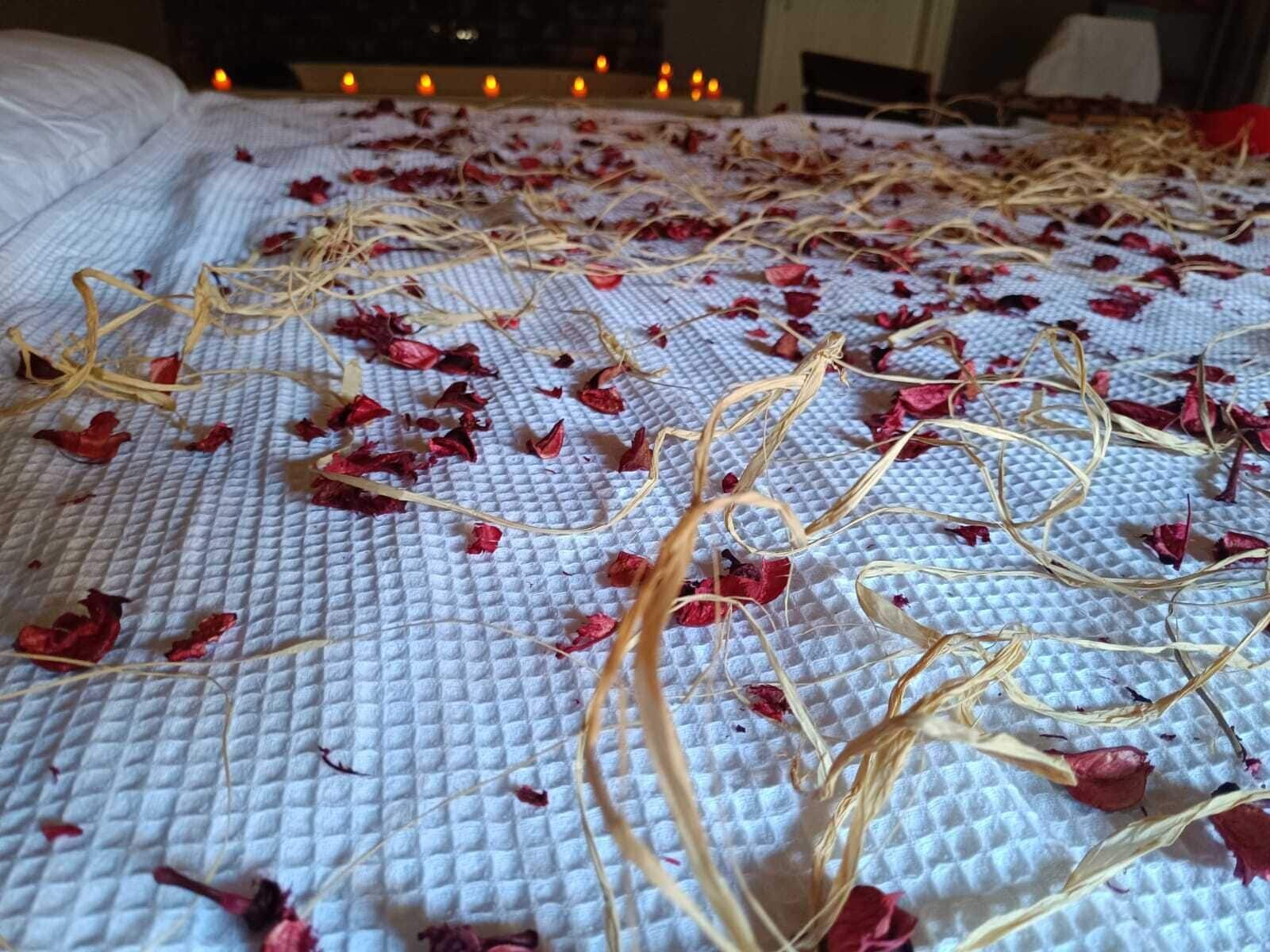 A bed with a textured white cover, scattered dried rose petals, and thin golden stems.