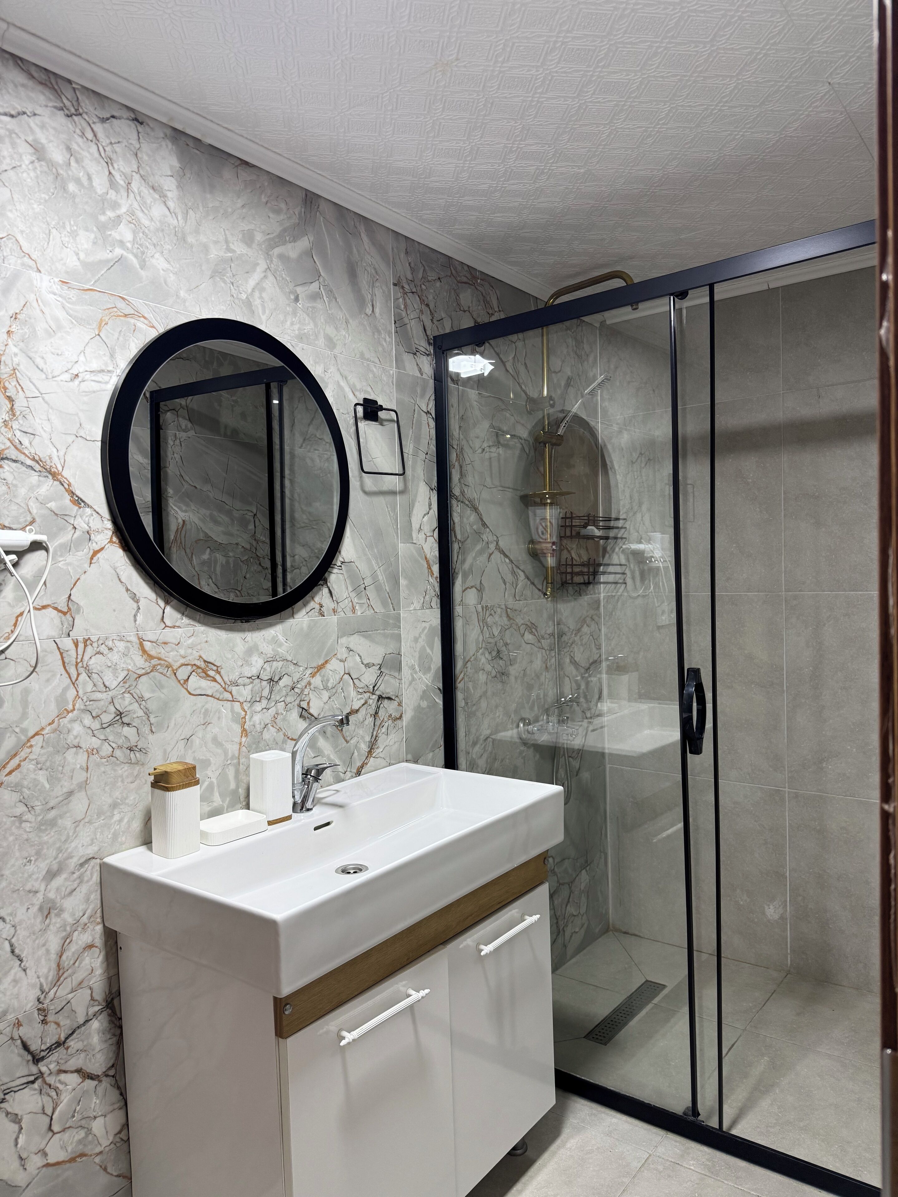 A modern bathroom with a marble backsplash, a round mirror, a white sink with a wooden countertop, and a glass-enclosed shower area.