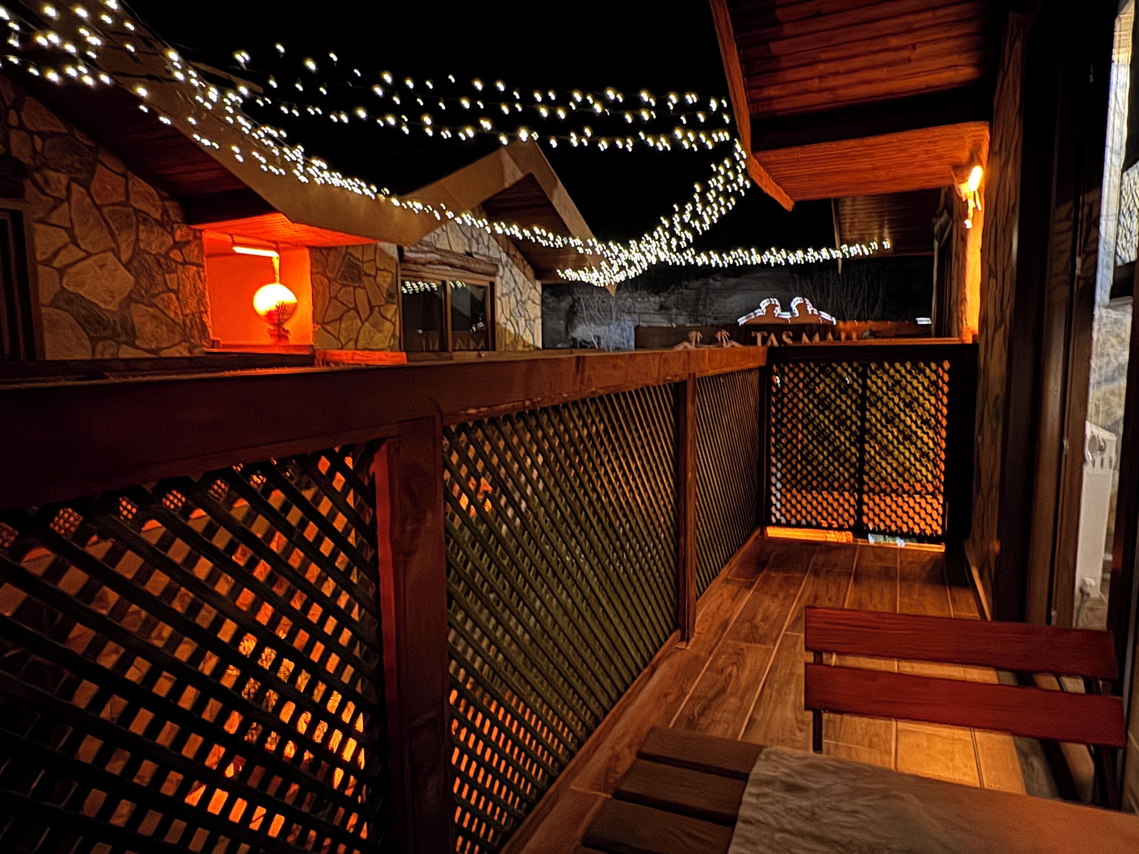 A wooden deck with a stone wall and a wooden railing. There are string lights above the deck.