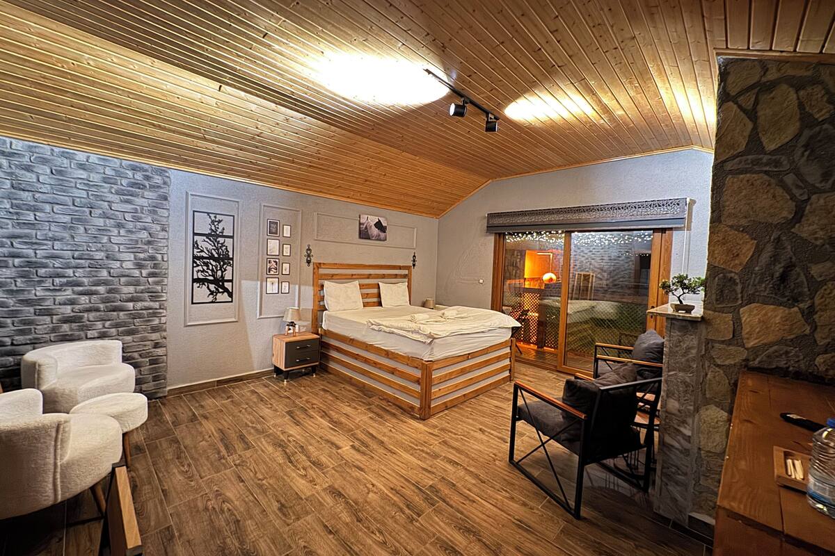 A bedroom with a wooden ceiling, a stone wall, a bed, a desk, and a chair.