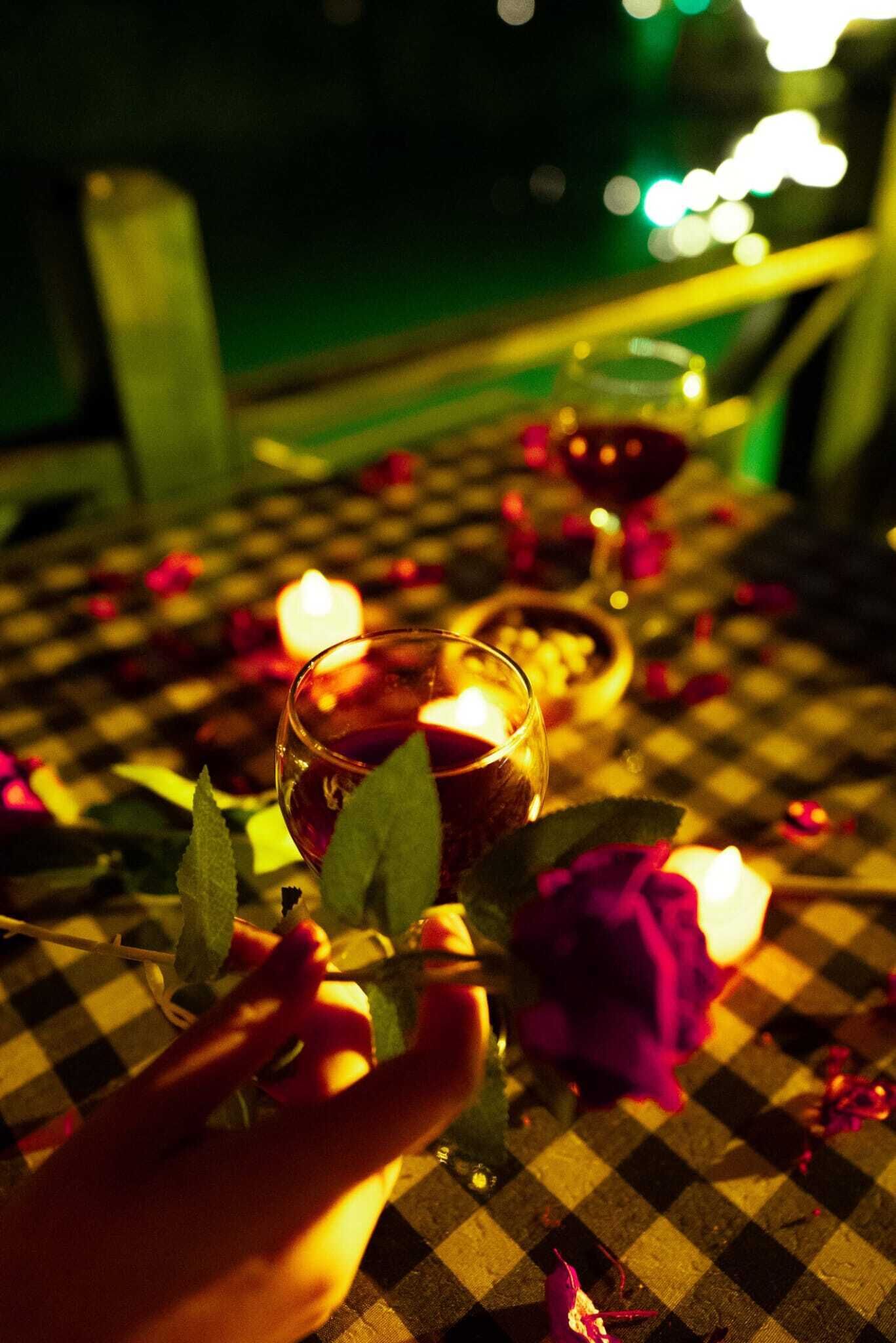 Uma mesa com toalha xadrez, um copo de vinho tinto, uma rosa e uma vela acesa.