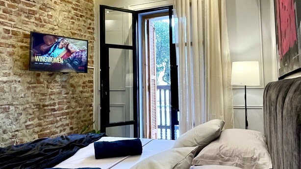 A modern hotel room with a brick wall, a television, a bed with pillows, a window with curtains, and a ceiling with recessed lighting.