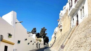 A narrow street flanked by white buildings with balconies, a clear blue sky, and a stone wall on one side.