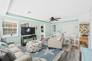 A living room with a television, a beige sofa, a round ottoman, and a ceiling fan.