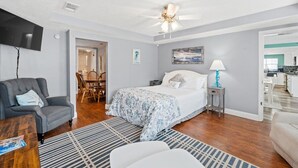 A bedroom with a bed, a chair, a TV, a ceiling fan, and a view into the kitchen. - Beach Better | Private Beach Access (Panama City Beach)
