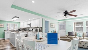 Eine moderne Küche mit weißen Schränken, eine Essecke mit weißem Tisch und Stühlen sowie ein Wohnzimmer mit einem beigen Sofa.