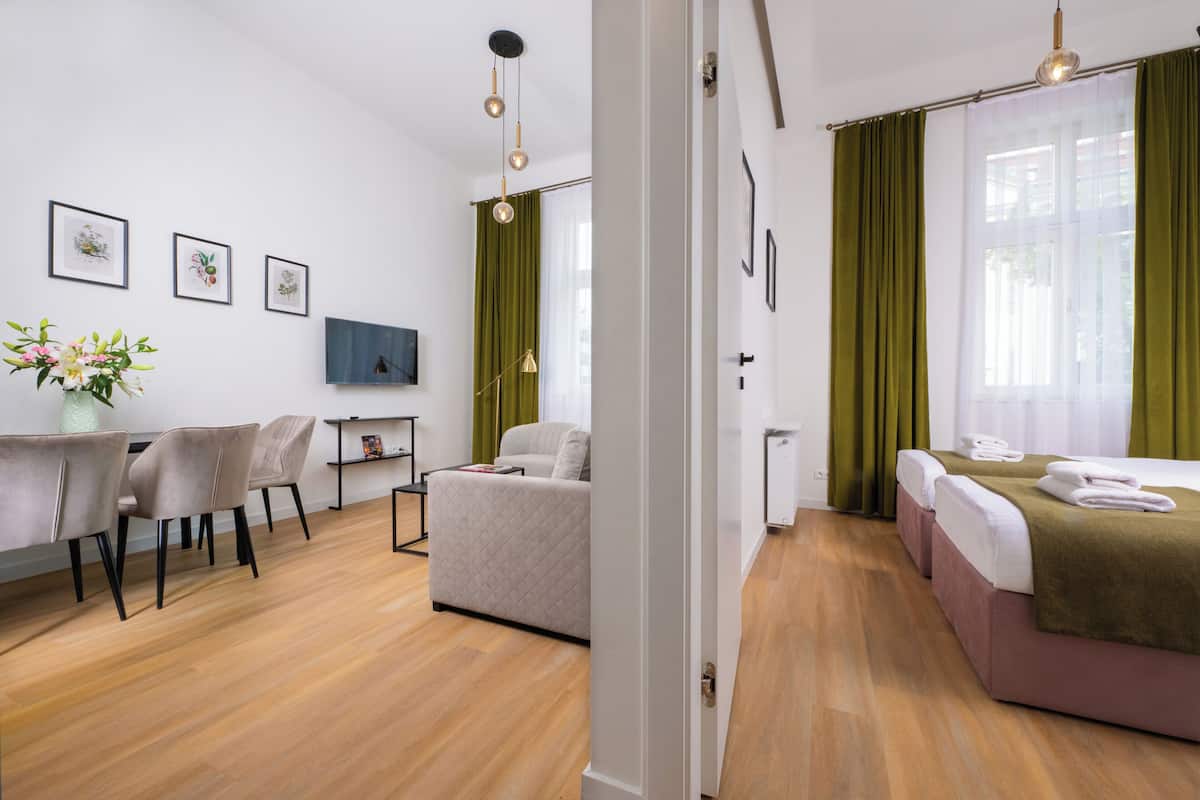 A modern hotel room with a sofa, a dining table with chairs, a TV, and a bed with green curtains.