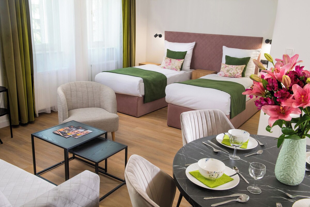 A hotel room with two beds, a sofa, a dining table set for two, and a vase of flowers.