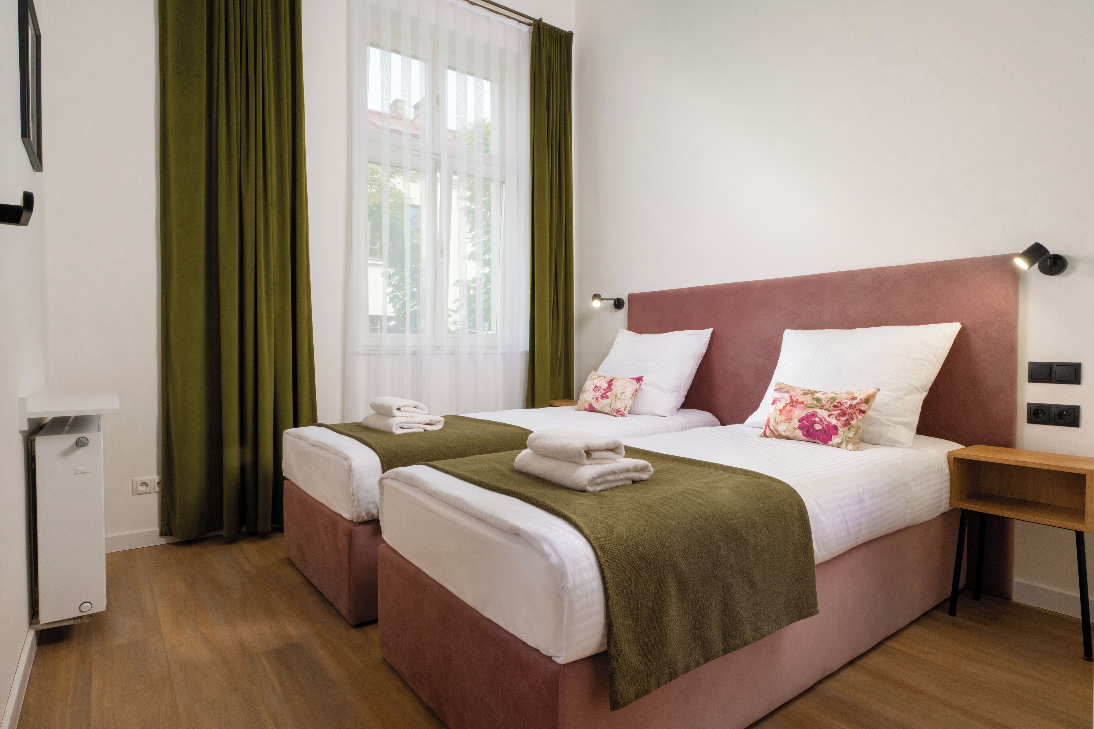 Ein Hotelzimmer mit zwei Betten, Holzboden, einem Fenster mit grünen Vorhängen und einem Nachttisch mit Lampe.