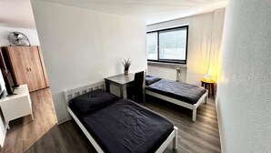 A compact bedroom with two single beds, a small table, a chair, a wardrobe, and a window with blinds.