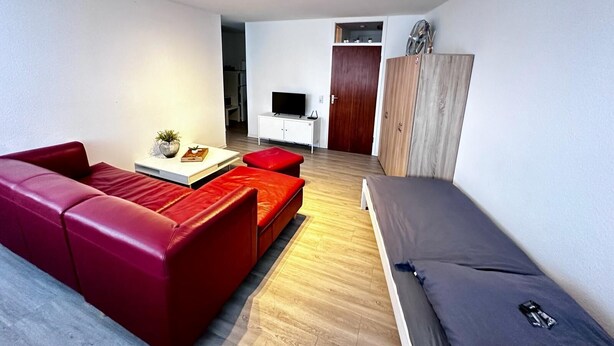 A modern living room with a red sofa, a small coffee table, and a grey sofa. There is a TV on a white cabinet, a wooden wardrobe, and a door leading to another room.