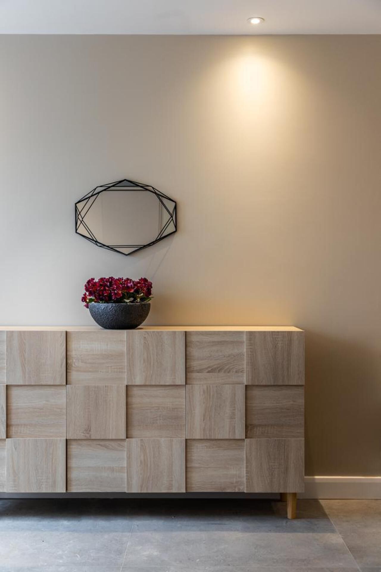 Un moderno mostrador de recepción con detalles de madera de diseño geométrico, un espejo decorativo y una planta con flores rojas.