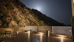 Um pátio externo com piso de pedra, cerca de madeira e vista para uma montanha sob céu noturno com uma lua visível.